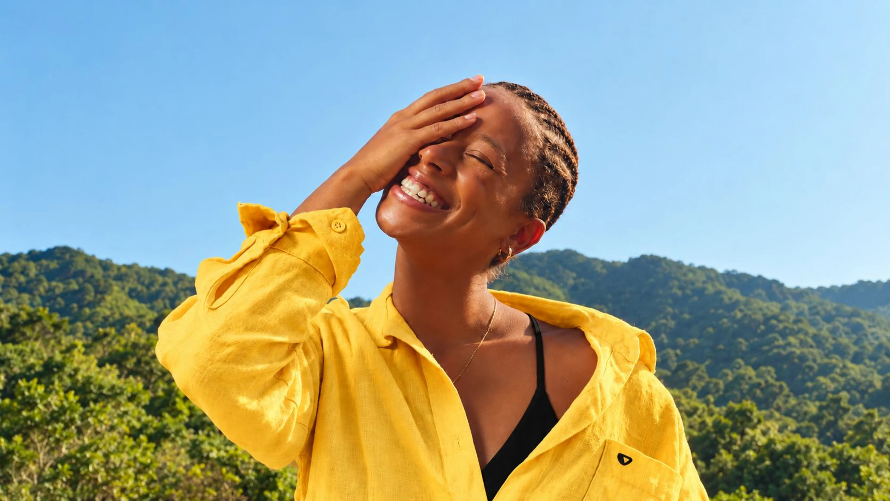 A young woman in a yellow shirt laughing in the sun, face turned up to an open blue sky, green hills behind